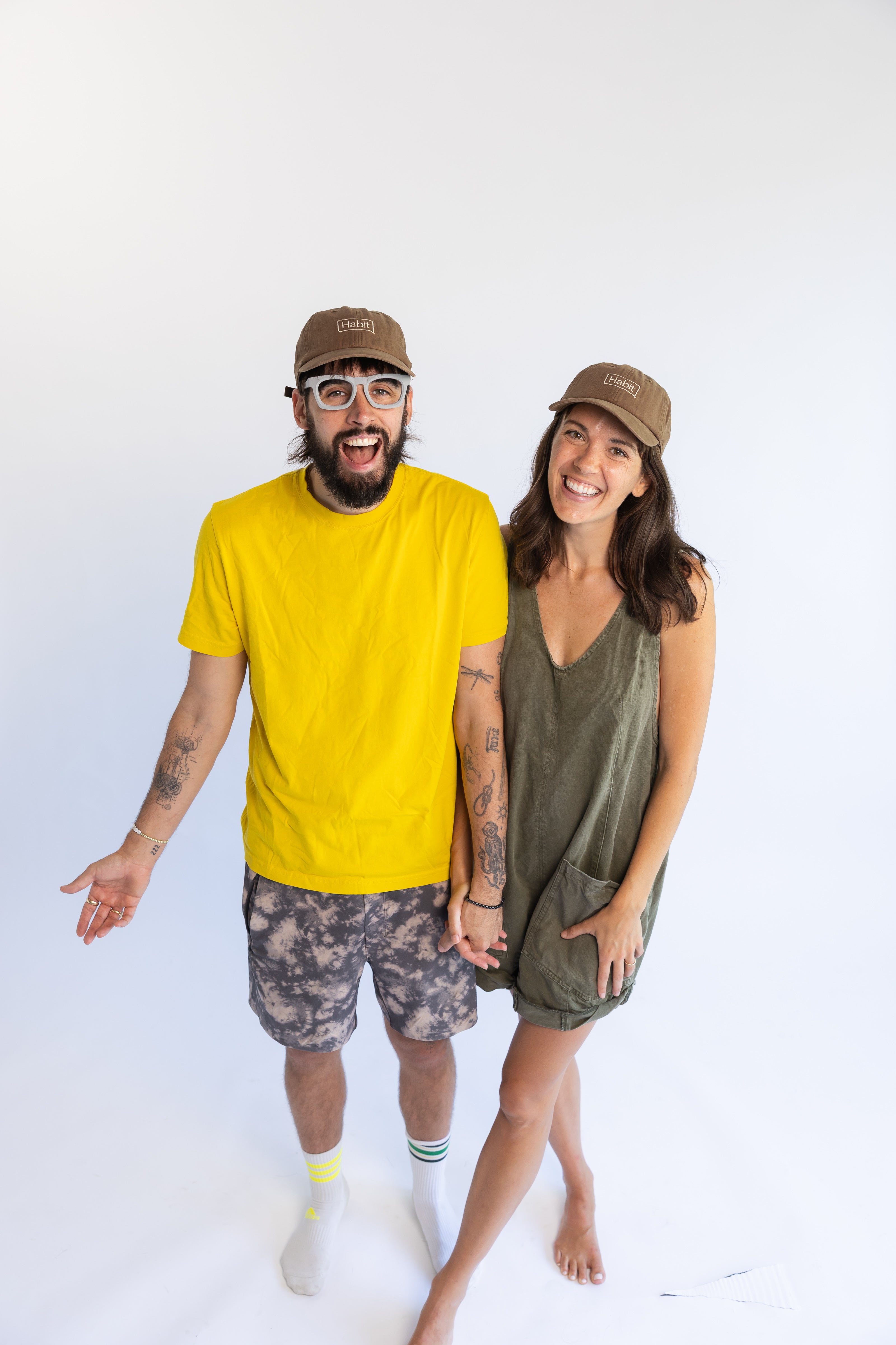 Two people wearing matching Habit hats, man in yellow shirt, woman in green shirt wearing baseball caps on a white background, Eamon and Bec