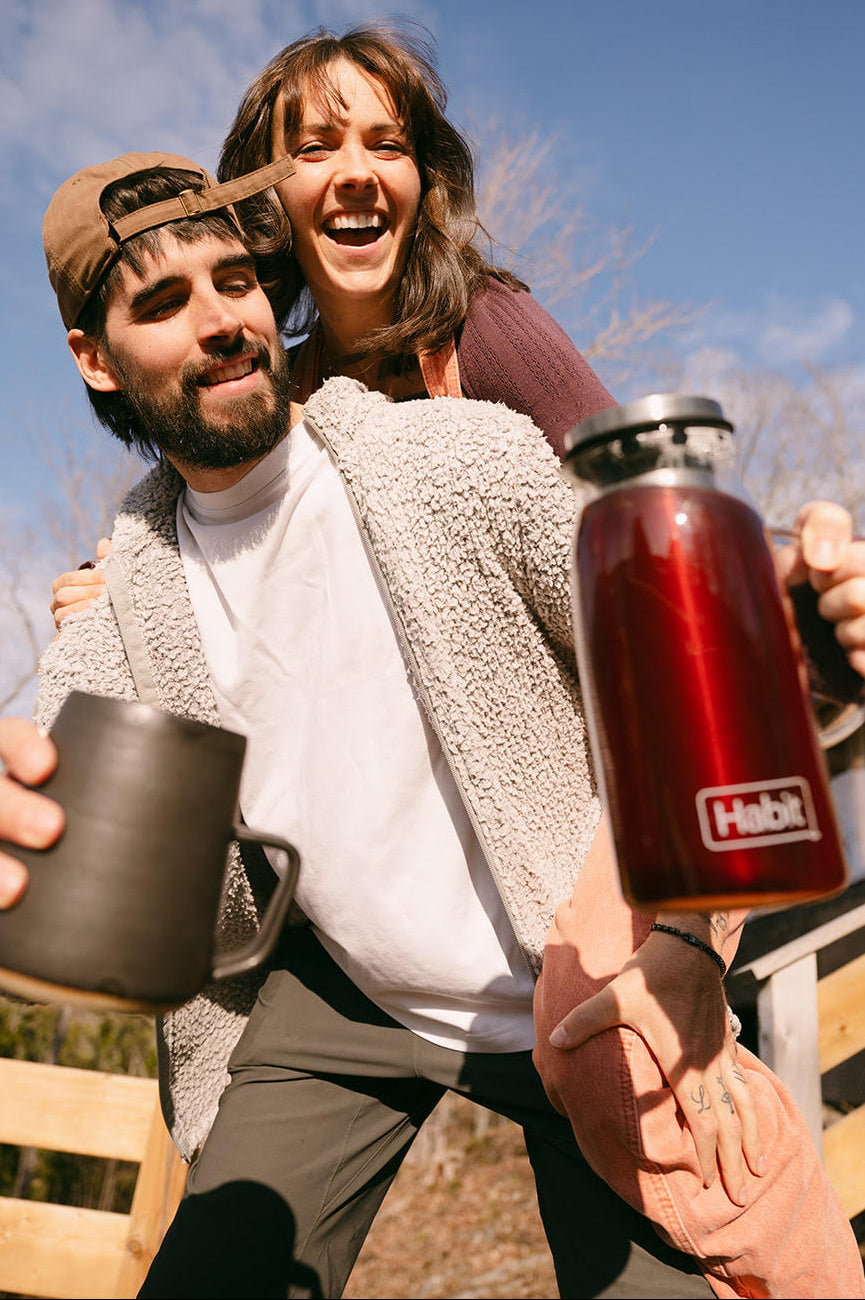 Two people outdoors, one holding a red insulated bottle and the other a black mug, with a clear blue sky in the background.