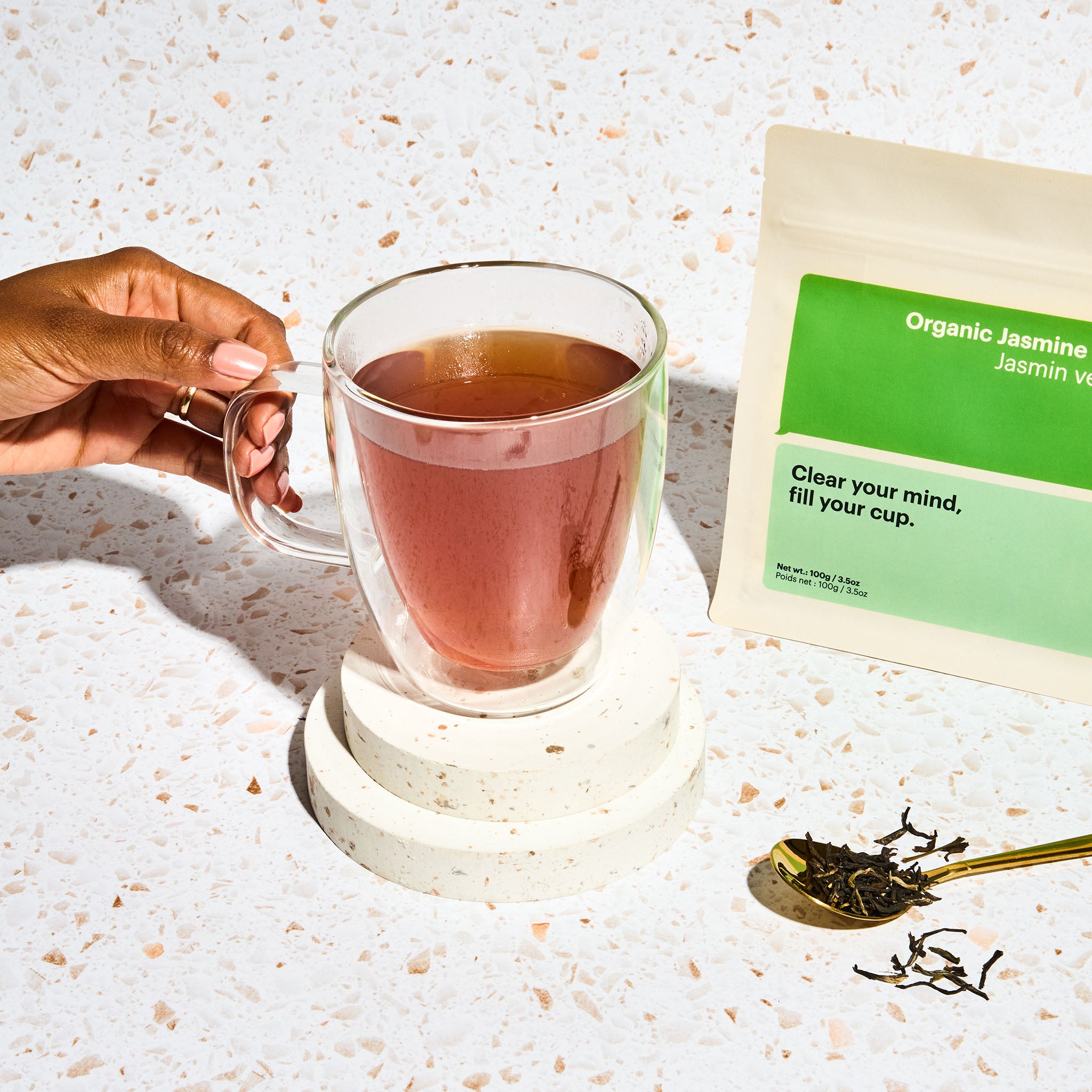 Cup of fresh brewed Organic Jasmine Green Tea with a woman's hand holding the cup, bag of tea in the background and a spoonful of loose leaf tea leaves next to the cup